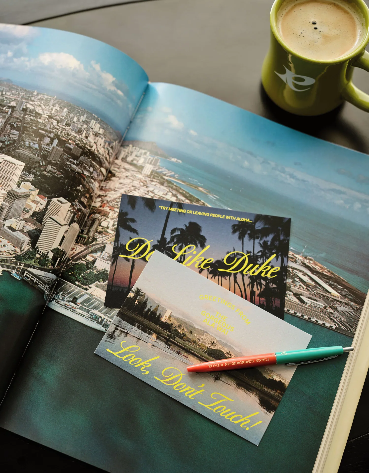 Standard room tabletop at Romer House Waikiki with open photo book, postcards, pen, and coffee mug.