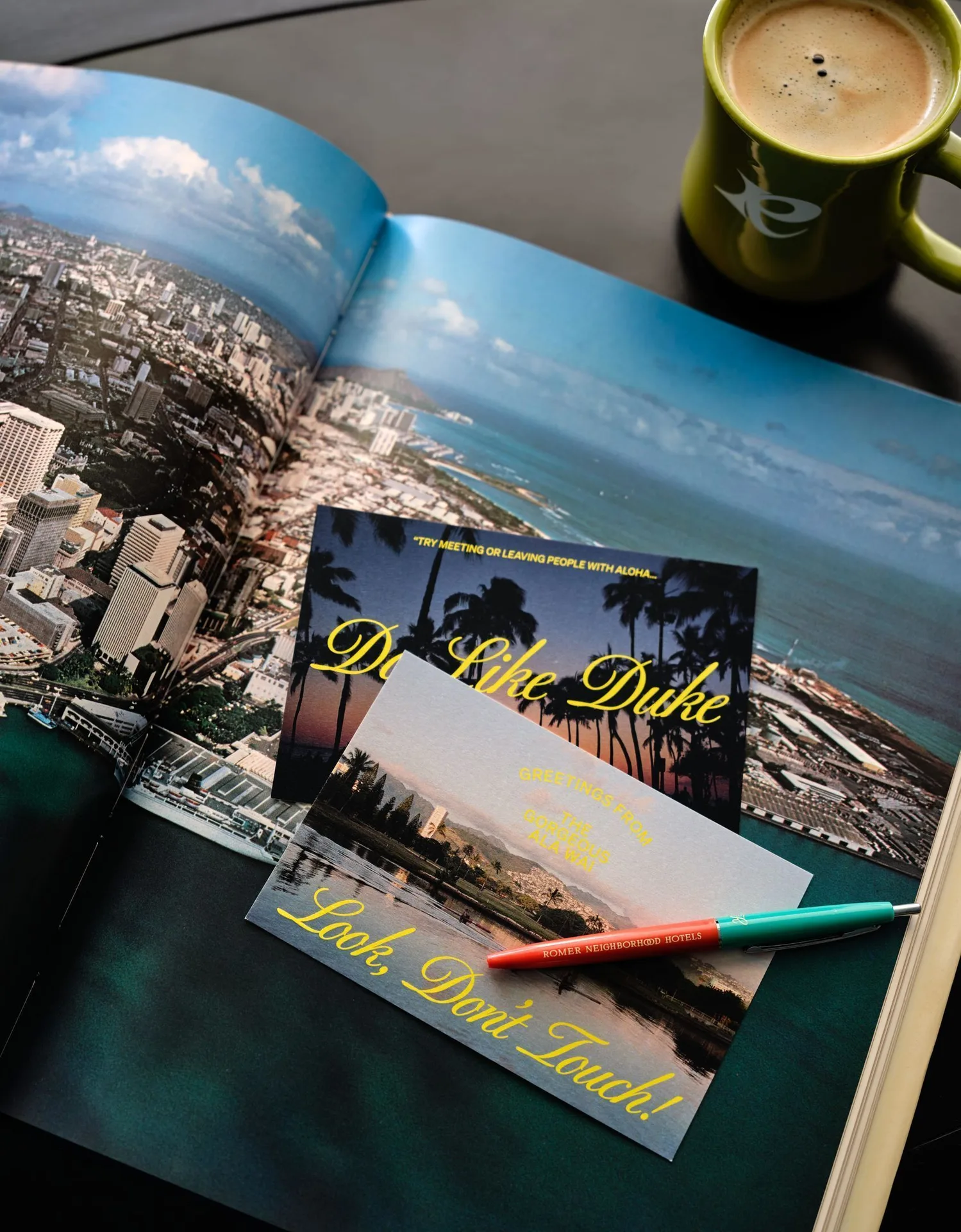 Standard Room tabletop with a Waikiki photo book, postcards, pen, and coffee at Romer House Waikiki.