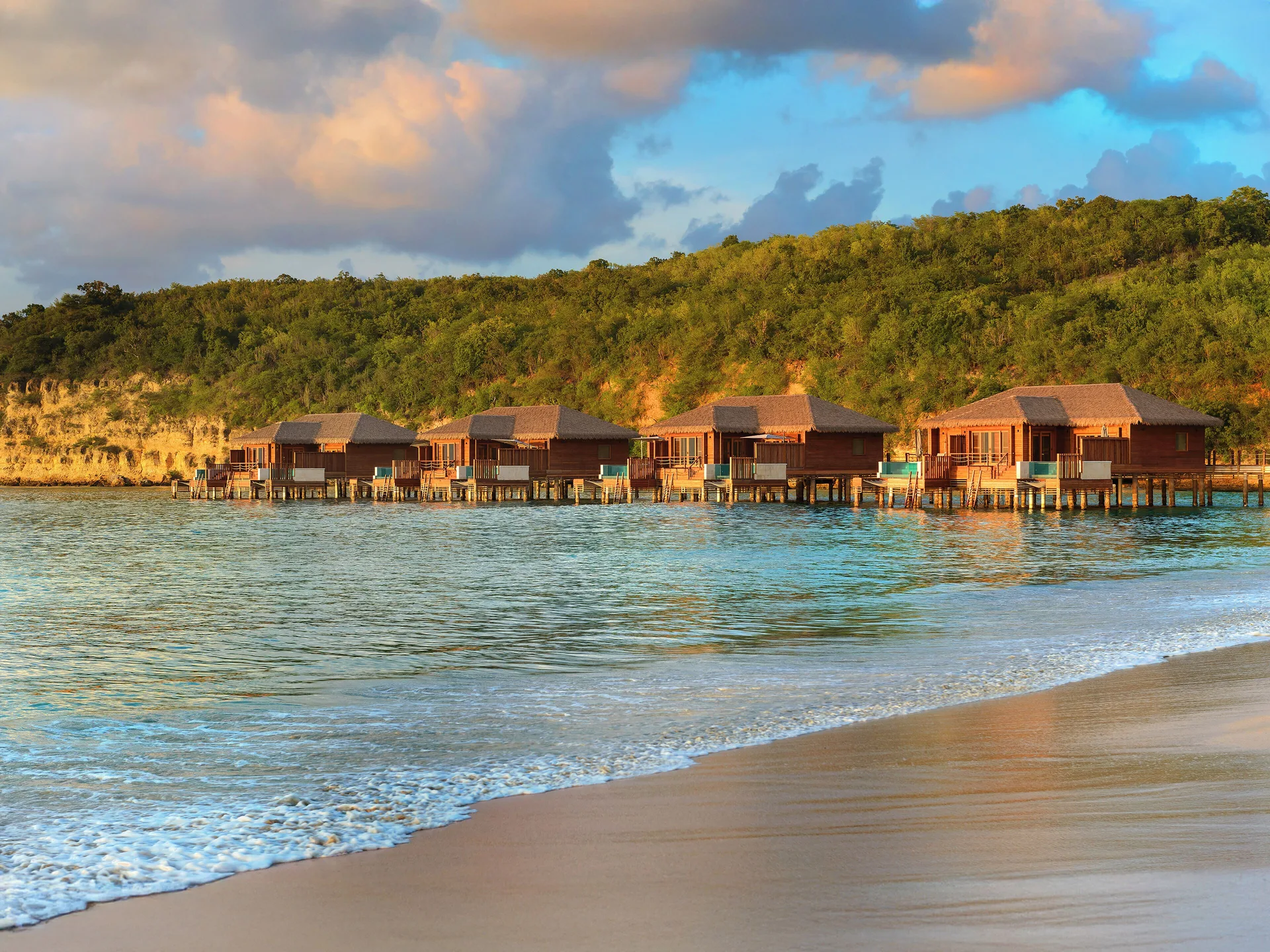 Overwater suites at Royalton CHIC Antigua viewed from the beach with waves in the foreground.