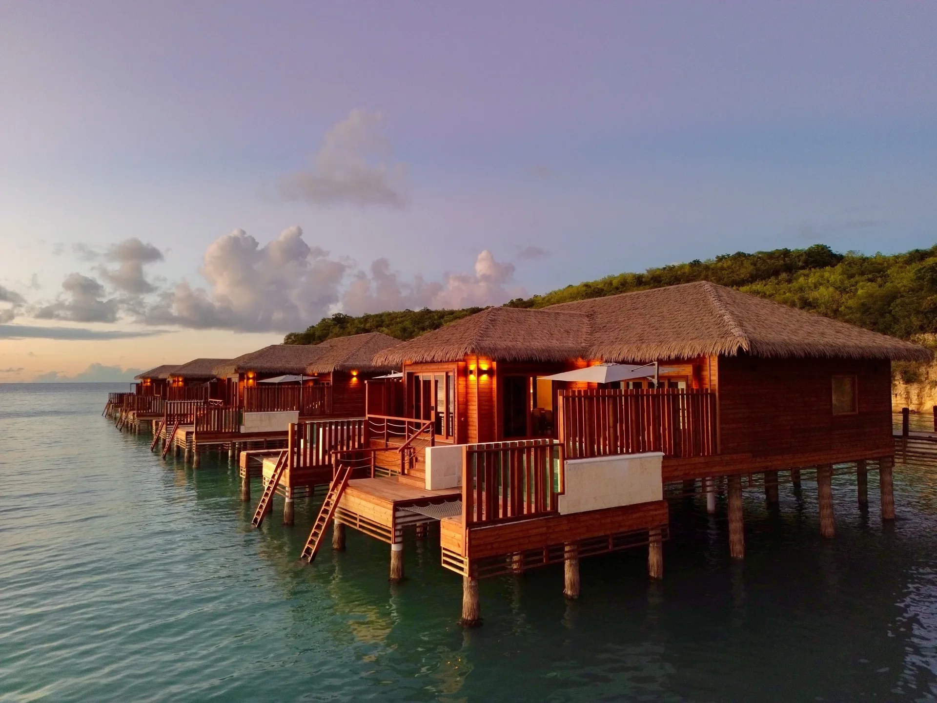 Thatched overwater suites at Royalton CHIC Antigua glowing at sunset above calm water.