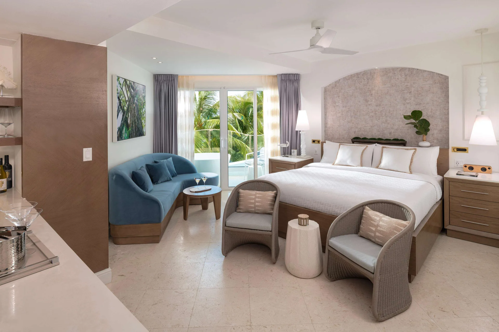 Guest room with king bed, blue sofa, chairs, bar area, and balcony doors at Sandals Dunns River.