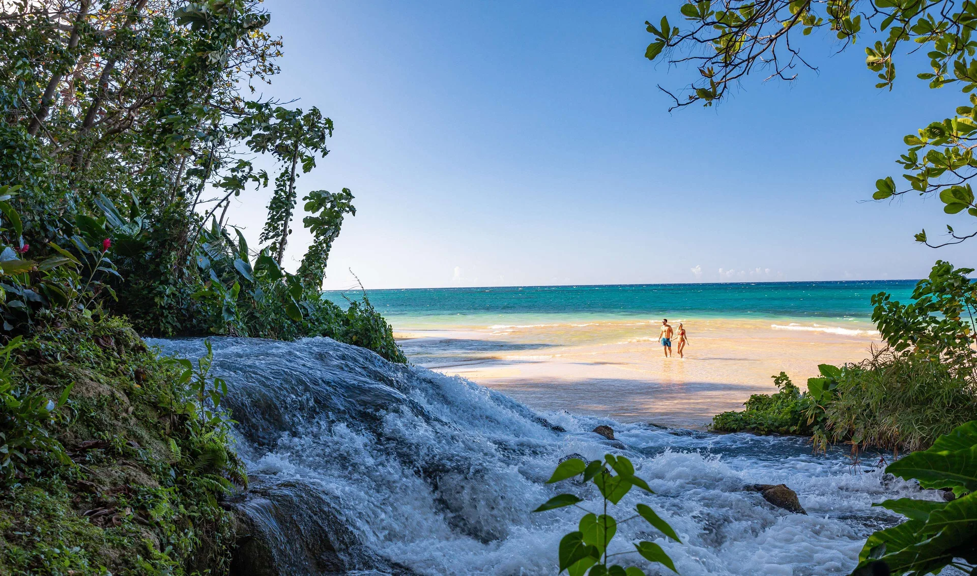 A waterfall frames two people walking on the beach at Sandals Dunns River.