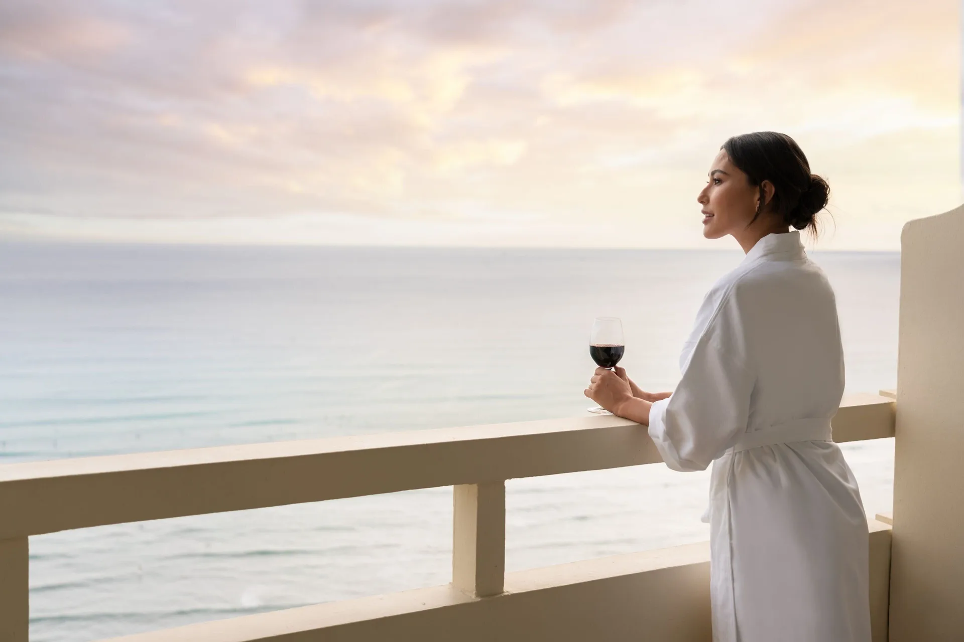 Guest in a robe holding a drink on an ocean-view balcony at Sheraton Waikiki Beach Resort.