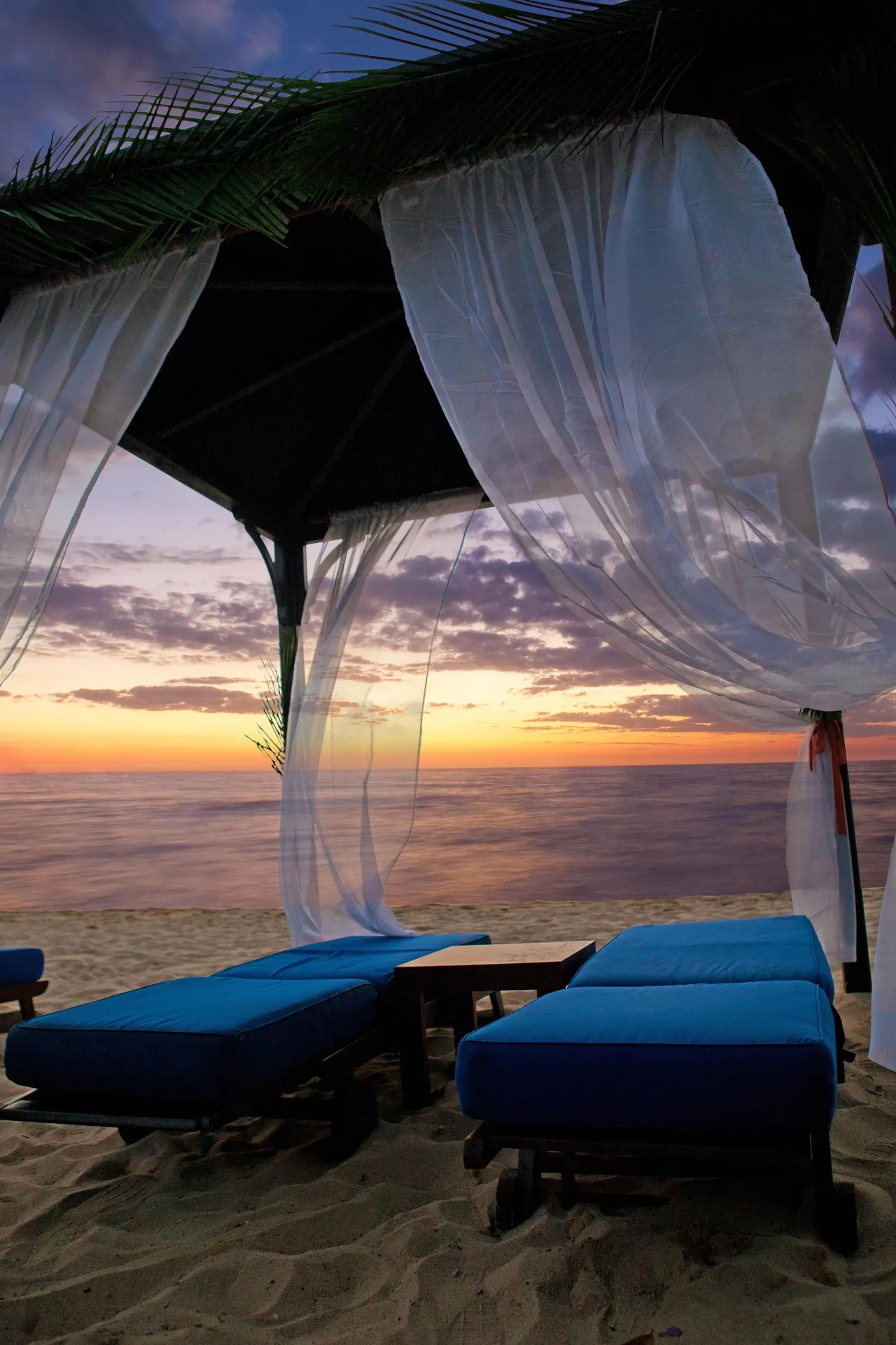 Beach cabana with blue loungers and sheer curtains at sunset at The House.
