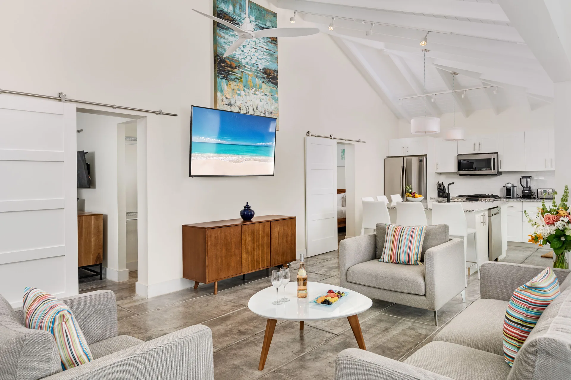 Villa living room with gray armchairs, round table, TV, and open kitchen at The Oasis at Grace Bay.