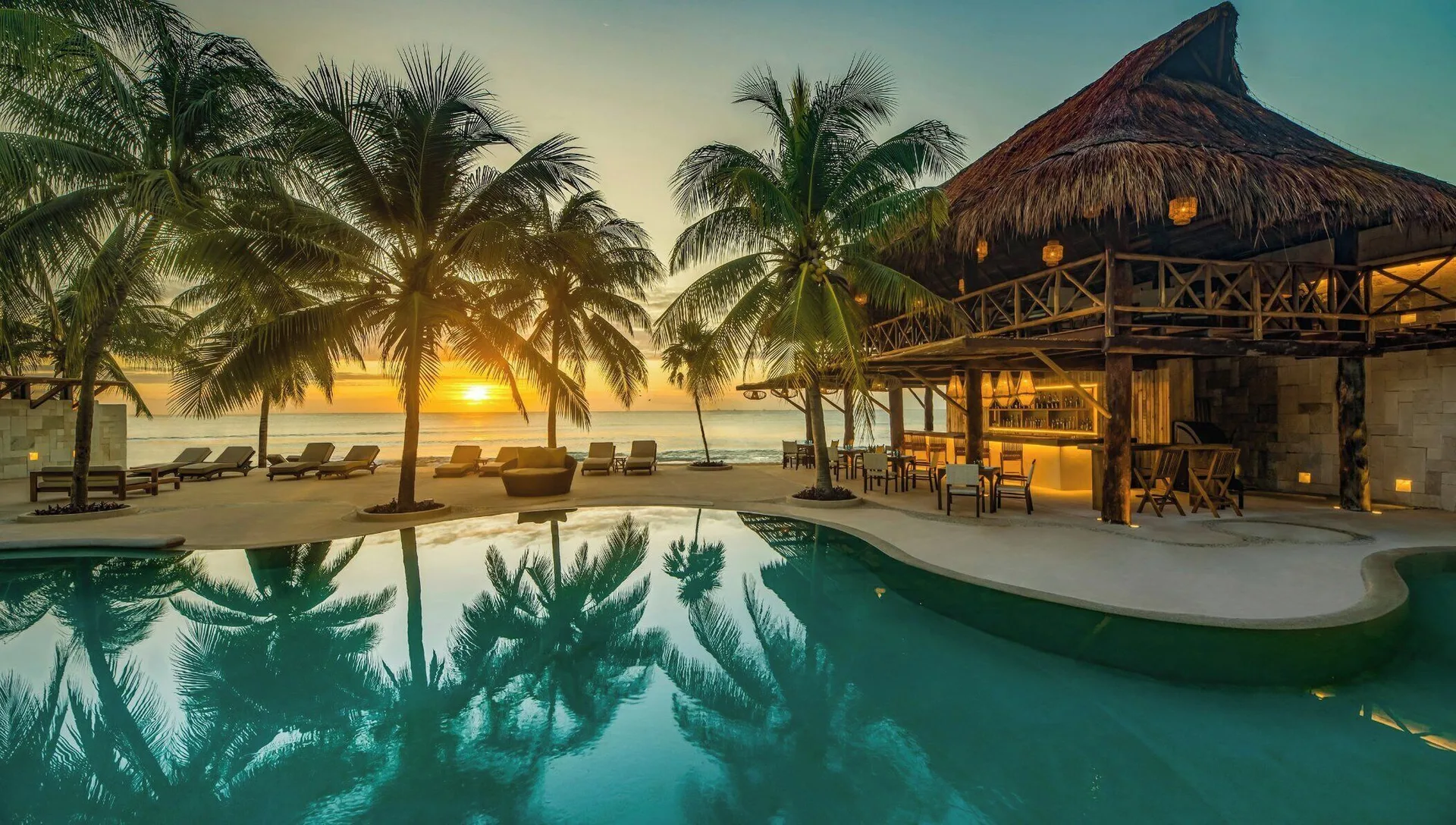 Beachfront pool, palm trees, and palapa building at sunset at Viceroy Riviera Maya.
