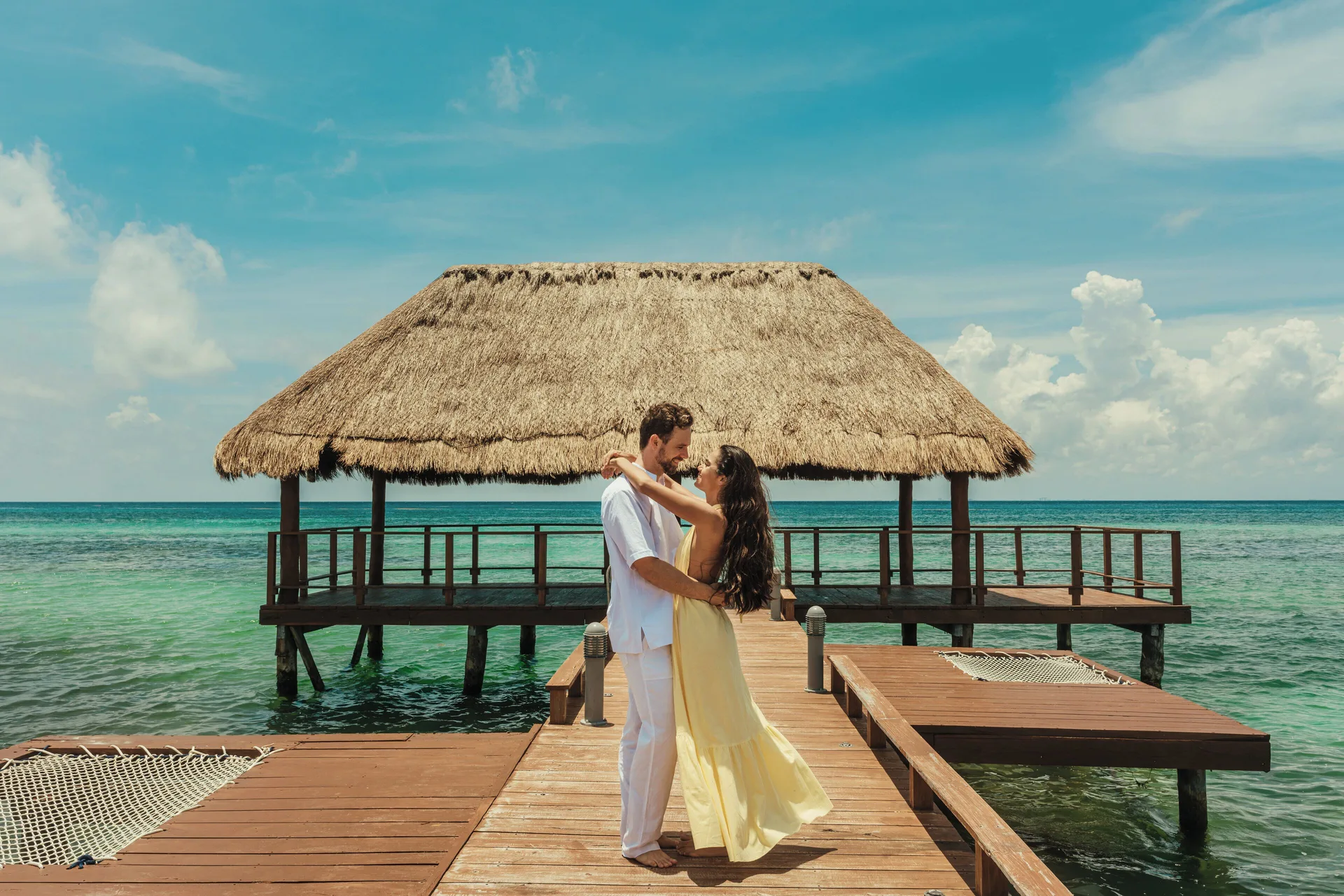 A couple stands on a wooden pier with a thatched pavilion over the sea at Viceroy Riviera Maya.